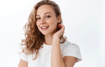 A person with curly hair smiles, wearing a white T-shirt, with a hand near their neck against a plain background.